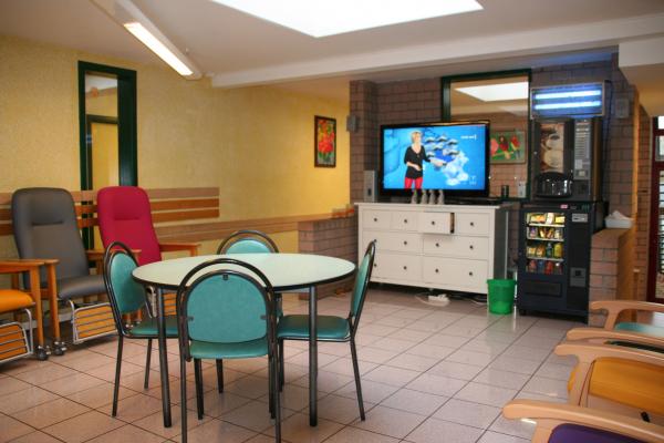 Le salon de la maison de repos en famille est est composé de fauteuils, de tables avec des chaises et une grande télévision. On voit aussi un distributeur de snacks et boissons