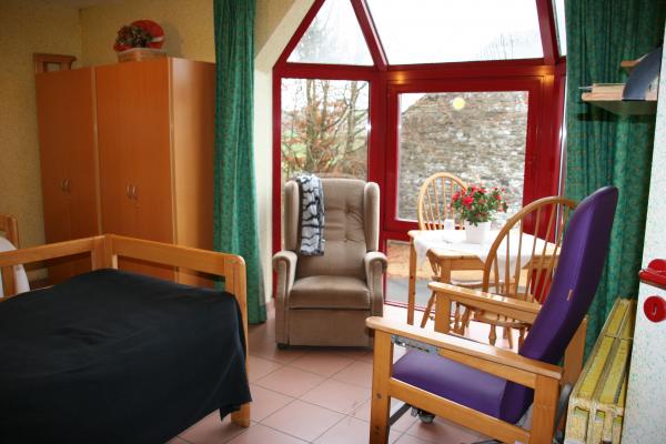 chambre de la maison de repos en famille avec un lit, des fauteuils, une table avec des fleurs, une fenêtre donnant sur un jardin et une grande armoire